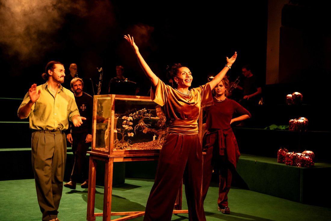 Eine Schauspielerin hebt begeistert die Arme. Dahinter schmunzeln sich eine junge Schauspielerin und ein junger Schauspieler belustigt zu. Zwischen den beiden steht eine Vitrine, darin ein Miniatur-Kirschbaum-Bonsai.