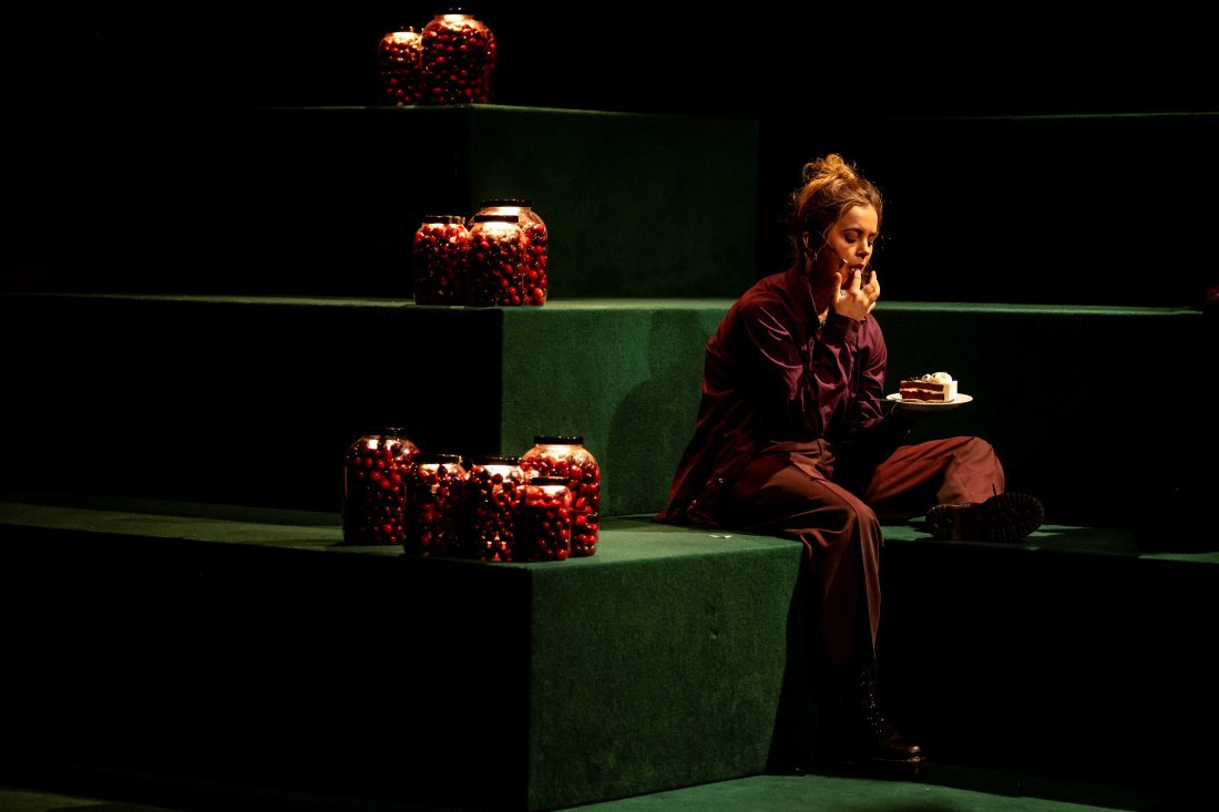 Eine junge Schauspielerin sitzt auf einer treppenartigen Bühne mit hohen Stufen, die mit grünem Filz ausgelegt ist. Nachdenklich ist sie ein Stück Schwarzwälder Kirschtorte.