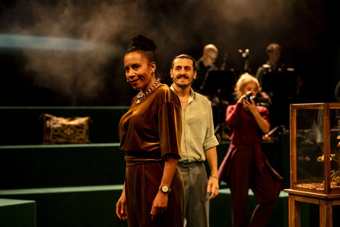 Drei Personen auch einer Bühne. Die erste, eine Frau mit goldenem Onesie, blickt verwegen ins Publikum, dahinter ein junger Mann der lacht und eine junge Frau, die beide filmt. Im Hintergrund Musiker*innen.