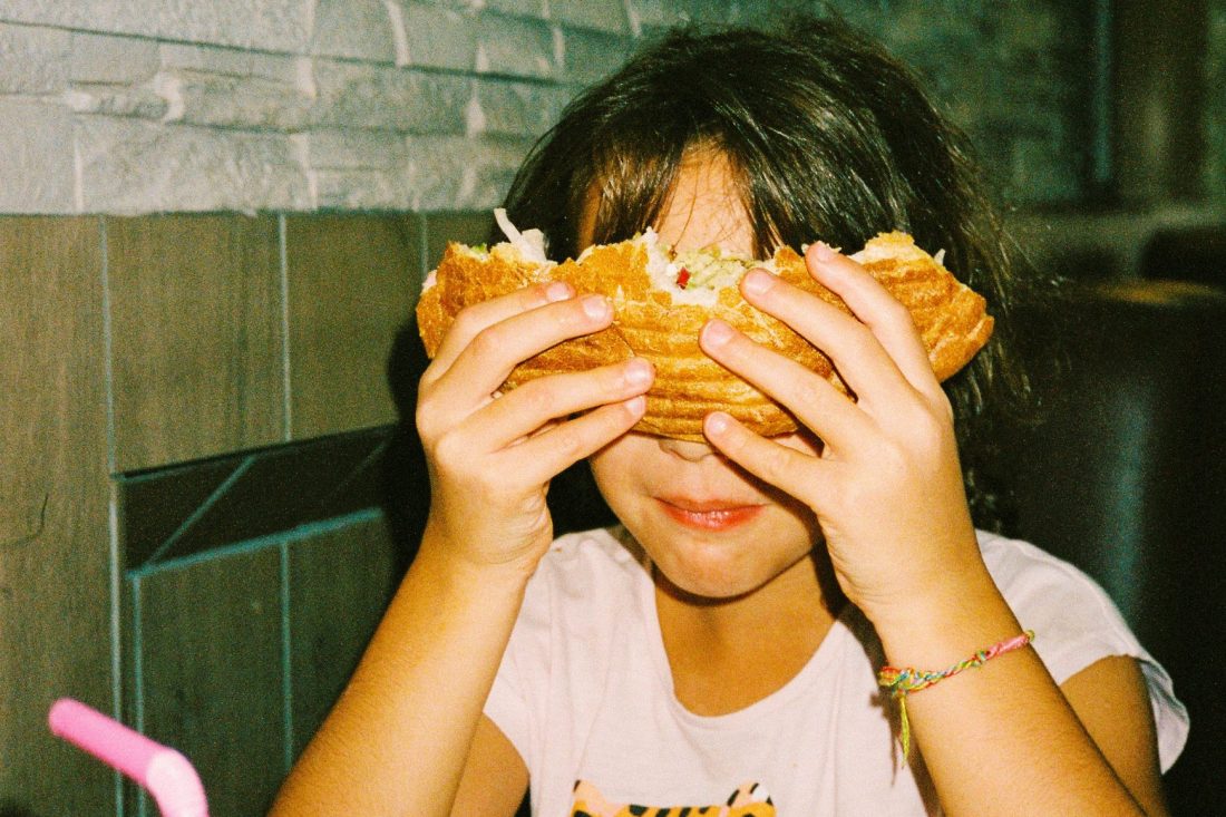 Ein junges dunkelhaariges Mädchen hält sich lächelnd einen angebissenen Döner vor das Gesicht.