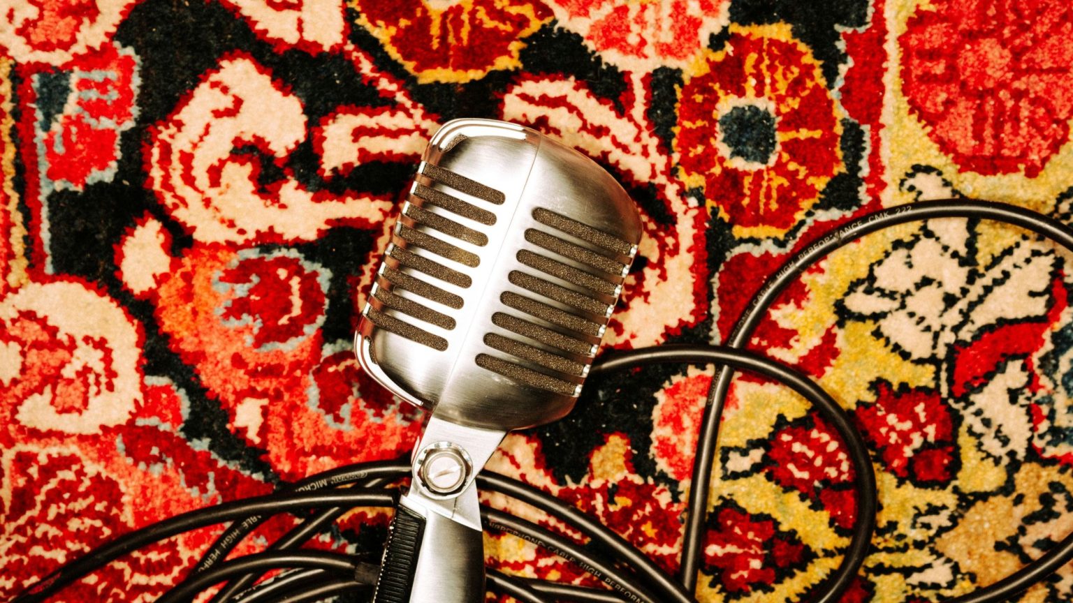 A silver vintage microphone lies on a bed of cables on a red, yellow, yellow, black, and white floral-patterned Persian carpet.
