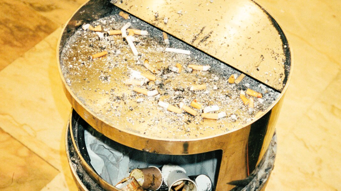 Ein vergoldeter Mülleimer, dessen Deckel als Aschenbecher voll ist mit Kippen. Darin liegen alte Verpackungen und Kaffeebecher, in denen auch ausgedrückte Kippen liegen.