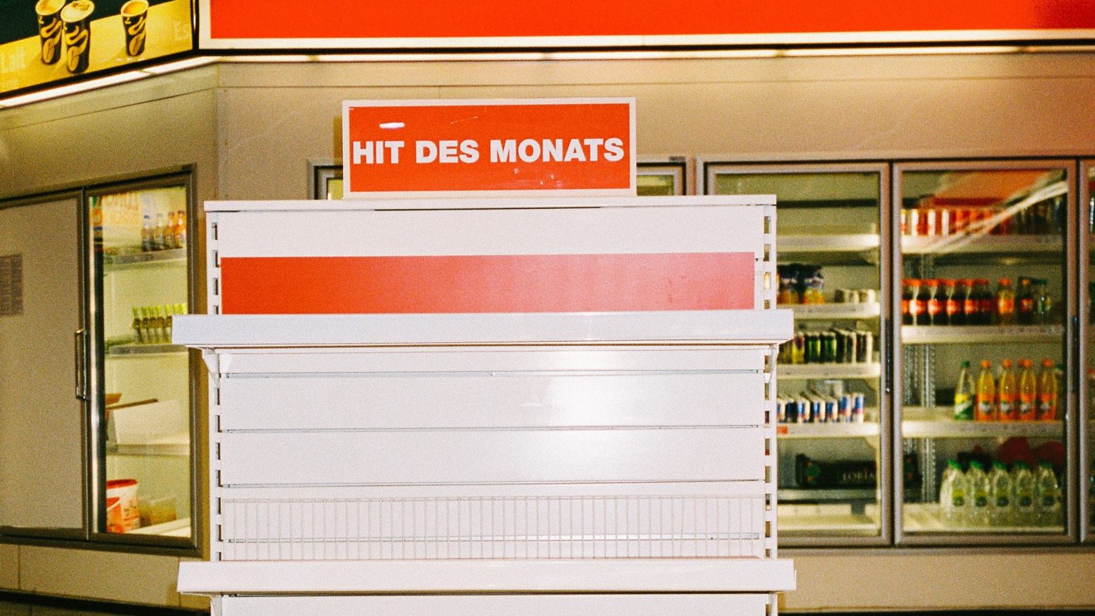 Eine Bahnhofshalle oder ein Kaufhaus im 90er-Stil. Im Vordergrund ein Stand, auf dem "HIT DES MONATS" steht, er ist allerdings leer und verbarrikadiert. Im Hintergrund Getränkeautomaten mit amerikanischen Softdrinks und ein Coffee to Go Shop.